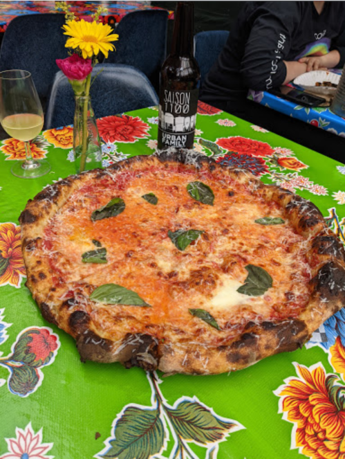 A large pizza topped with cheese and basil on a colorful floral tablecloth, with a beer bottle and glass nearby.