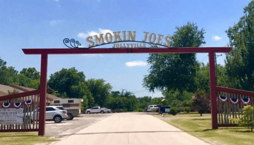 Sign for "Smokin Joe's" at the entrance of a venue, surrounded by trees and a clear blue sky.