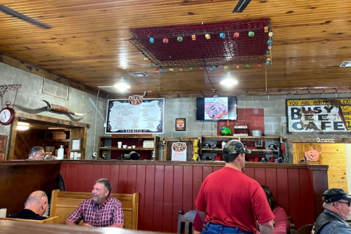 Interior of a rustic cafe with wooden booths, patrons dining, and a menu board on the wall. A red grid hangs from the ceiling.