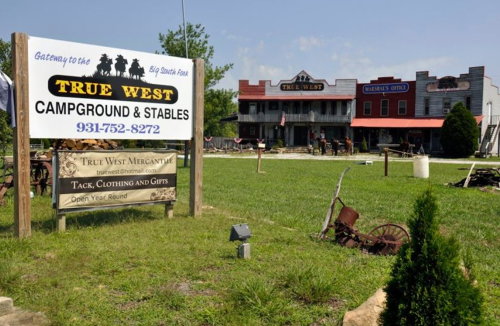 Sign for True West Campground & Stables with a western-themed building in the background, surrounded by green grass.