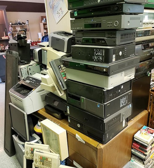 A cluttered room filled with stacked old electronics, including printers, computers, and various devices on a wooden table.