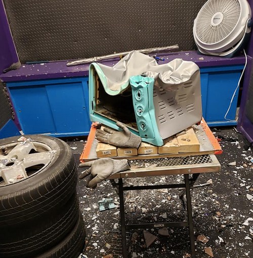 A damaged toaster on a table surrounded by debris, tires, and a fan in a chaotic indoor space.