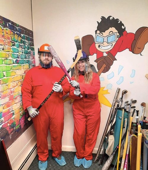 Two people in red jumpsuits and helmets hold hockey sticks, standing in front of a colorful mural of a cartoon character.