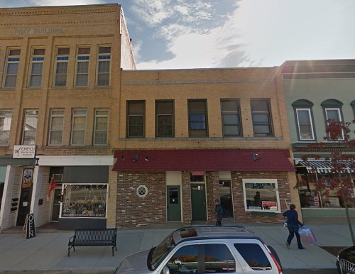 A street view of a brick building with multiple windows, a red awning, and nearby shops and a bench.