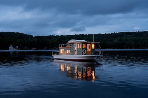 A houseboat illuminated at dusk, floating on calm water with trees in the background.