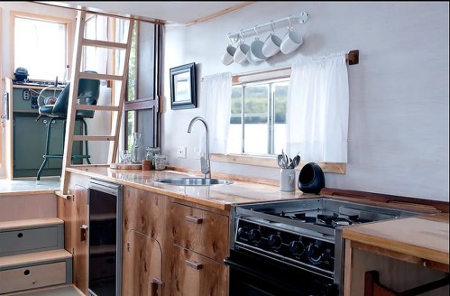A cozy kitchen with wooden cabinets, a sink, stove, and a ladder leading to a loft area with a view.