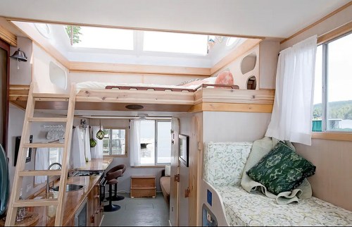 Cozy tiny home interior featuring a lofted bed, kitchenette, and seating area with large windows for natural light.