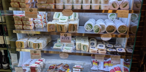 A display case filled with various types of cheese, labeled with names and descriptions, alongside bottles of wine.