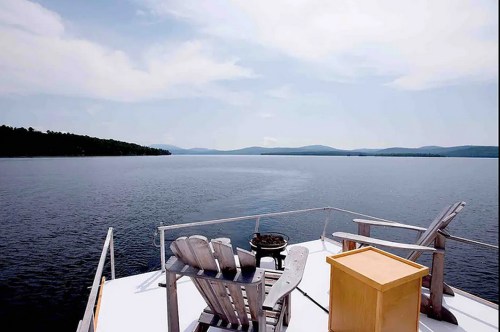A serene lakeside view from a deck with two chairs and a small table, surrounded by mountains under a cloudy sky.