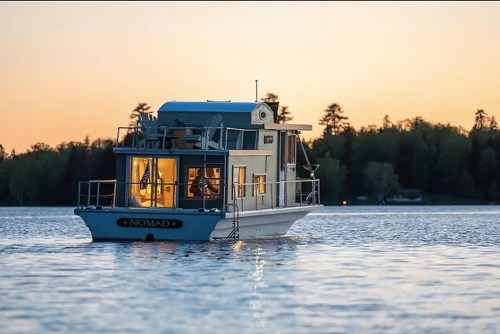 A houseboat named "Nomad" floats on calm water at sunset, surrounded by trees and a serene landscape.