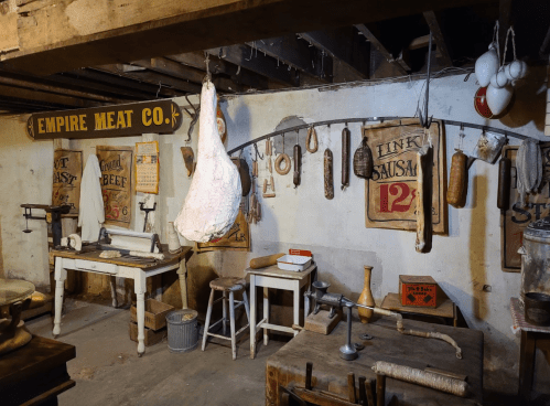 A rustic meat processing room with hanging meats, tools, and vintage signs from "Empire Meat Co." on the walls.