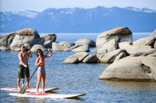 Two people paddleboarding on a calm lake surrounded by large rocks and mountains in the background.