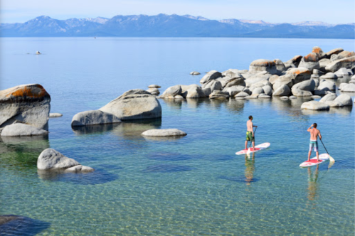 Two people paddleboarding on clear water surrounded by rocks, with mountains in the background.