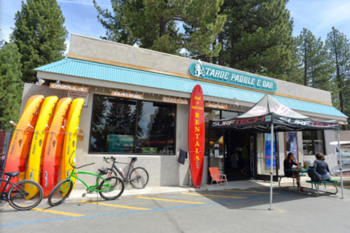 Exterior of Tahoe Paddle & Oar shop with colorful kayaks, bikes, and a rental tent under a blue sky and tall trees.