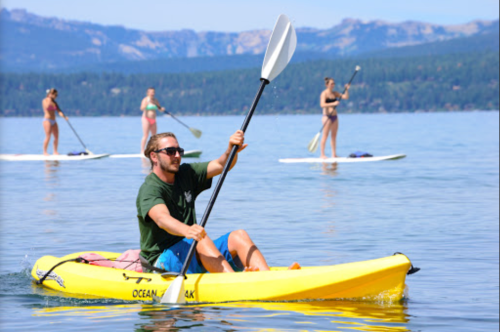 A man paddles a yellow kayak on a lake, with people paddleboarding in the background and mountains in the distance.