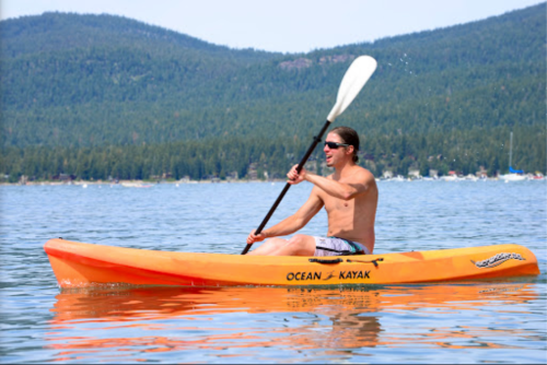 A person paddles a bright orange kayak on a calm lake surrounded by trees and mountains on a sunny day.