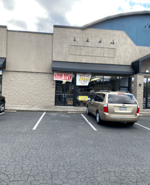 A storefront with a "NOW OPEN" sign, featuring a parked car in front and a cloudy sky above.