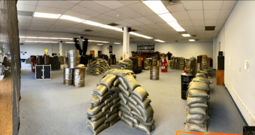 A spacious indoor arena filled with stacked sandbags, barrels, and obstacles for paintball or airsoft games.