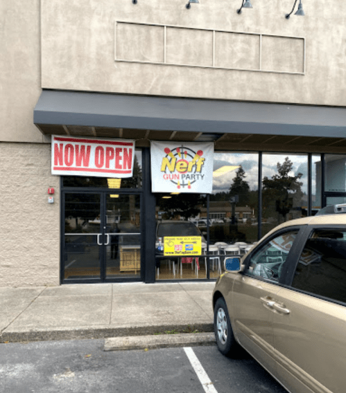 A storefront with a "Now Open" sign and a banner for a Nerf gun party. A car is parked in front.