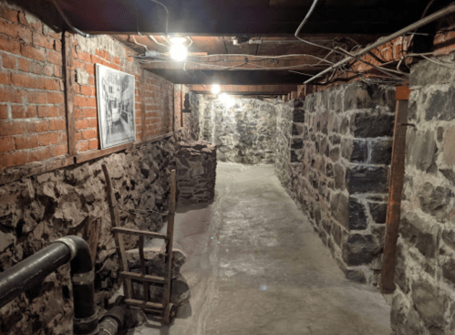 A dimly lit basement corridor with brick and stone walls, featuring a photo on the wall and a wooden chair.