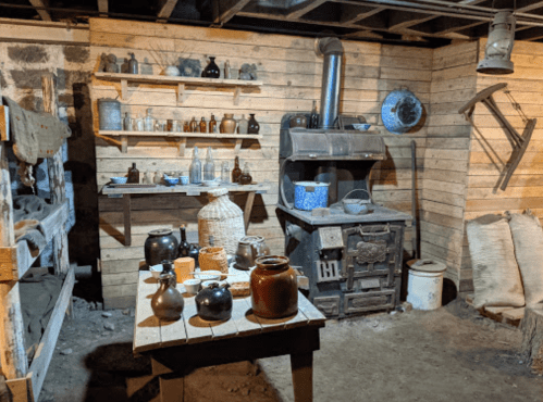 A rustic kitchen with wooden shelves, various jars, a stove, and a table filled with pottery and woven baskets.