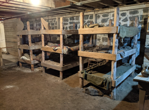 A dimly lit basement with wooden bunk beds and simple bedding, set against a stone wall.