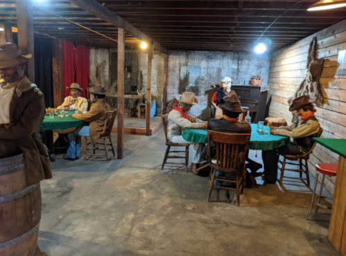 A dimly lit room with several cowboys seated at green poker tables, surrounded by rustic decor and wooden barrels.