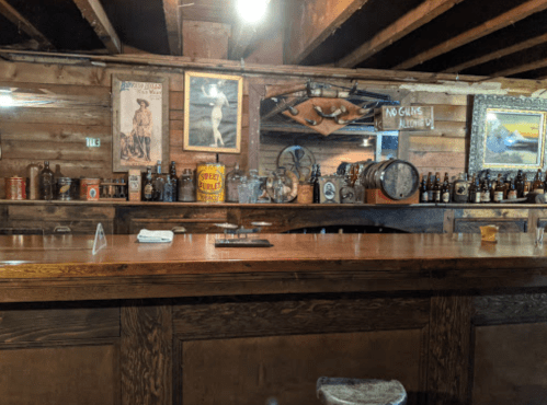 A rustic bar with wooden beams, vintage decor, and various bottles displayed on shelves. A sign reads "No guns allowed."