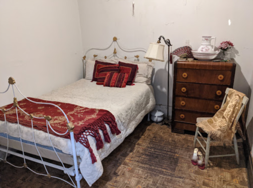 Cozy bedroom with a vintage bed, decorative pillows, a wooden dresser, and a chair beside a lamp.