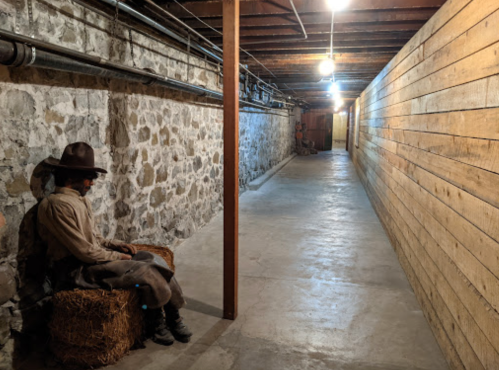 A dimly lit basement corridor with stone walls, wooden panels, and a figure sitting on a hay bale.