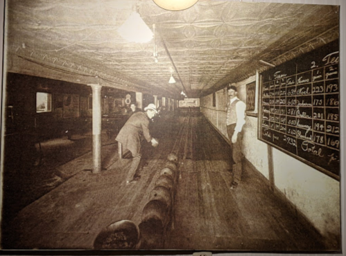 A vintage bowling alley with two men; one is rolling a ball while the other watches, with a scoreboard on the wall.