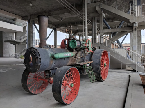 A vintage steam tractor with red wheels and green accents, displayed in a modern industrial setting.