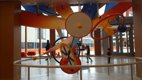 Colorful abstract sculpture in a spacious indoor area, with children exploring around it. Large windows in the background.
