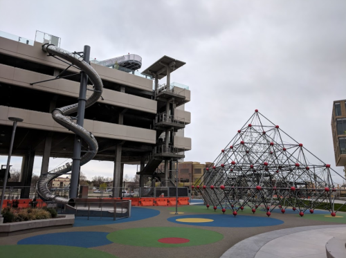 A modern playground featuring a slide, climbing structure, and colorful ground surfaces, set against a cloudy sky.