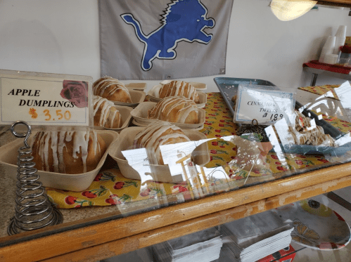 Display case with apple dumplings and cinnamon rolls, featuring price tags. A Detroit Lions flag is in the background.