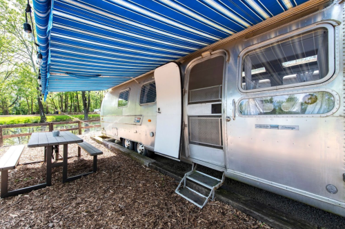 A shiny Airstream trailer with a blue striped awning, next to a picnic table in a wooded area.