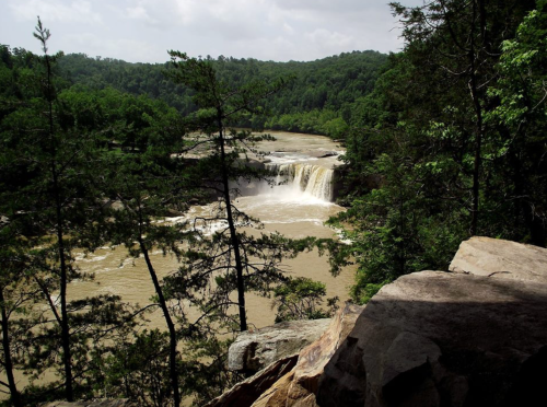 A scenic view of a waterfall surrounded by lush green trees and rocky terrain under a cloudy sky.