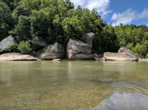 A serene river scene with large rocks and lush green trees lining the banks under a partly cloudy sky.