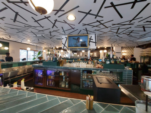 A modern bar with a green counter, shelves of drinks, and a patterned ceiling, bustling with staff and patrons.