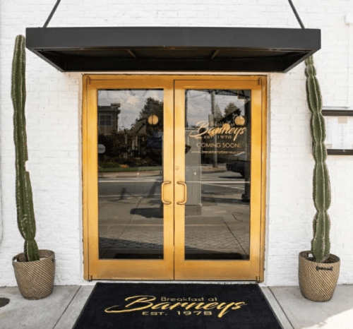 Entrance to Barneys, featuring golden double doors, a "Coming Soon" sign, and tall cacti in decorative pots.