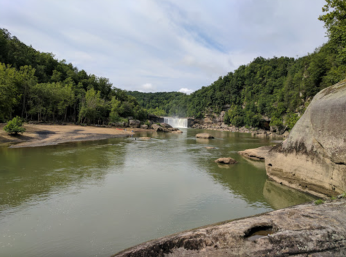 A serene river scene with a waterfall in the background, surrounded by lush green trees and rocky formations.