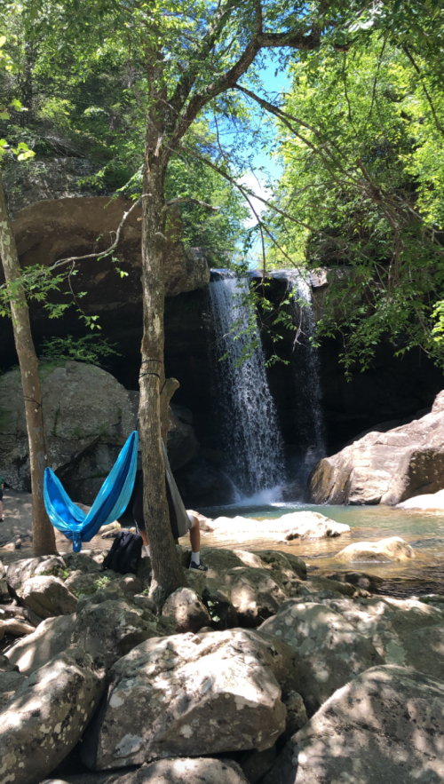 A serene waterfall cascades into a rocky stream, with a hammock set up between trees nearby.