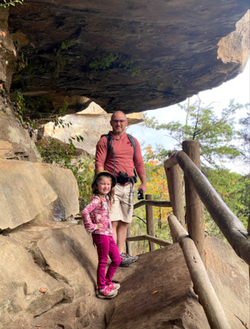 A man and a young girl stand on a rocky trail under a large overhang, surrounded by trees and autumn foliage.
