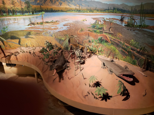 Diorama depicting prehistoric landscape with dinosaurs and aquatic reptiles near a river, surrounded by lush vegetation.