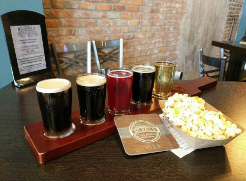 A flight of five craft beers on a wooden tray next to a serving of popcorn, with a brick wall in the background.