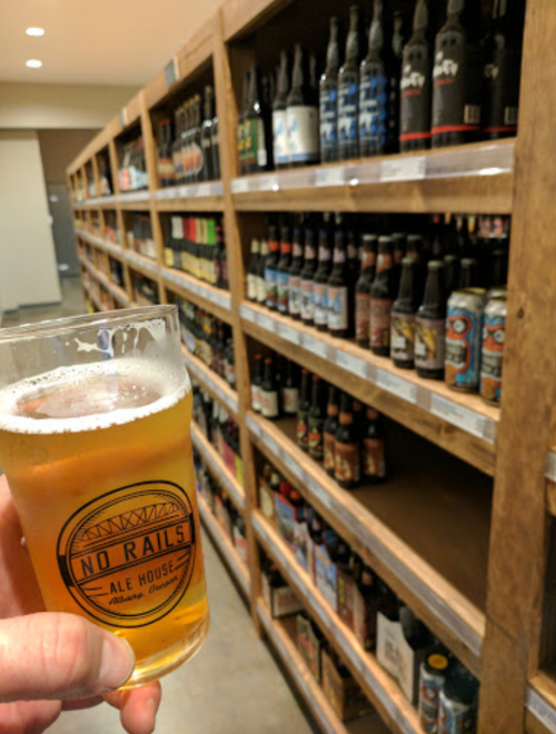 A hand holding a glass of beer in front of shelves filled with various beer bottles.