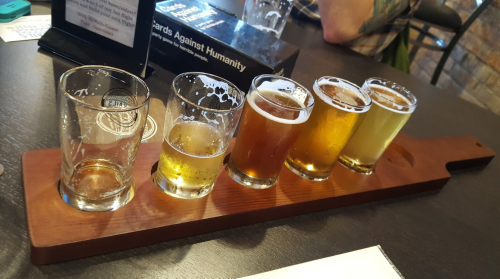 A wooden flight board with five small glasses of different beers, some partially empty, on a table.