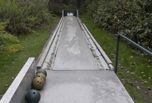 A bowling lane outdoors with three bowling balls in the foreground and pins at the end, surrounded by greenery.