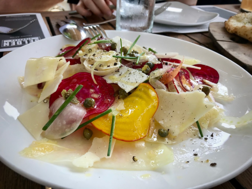 A colorful salad with sliced beets, cheese, capers, and herbs on a white plate, served on a wooden table.