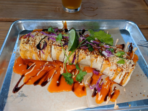 A plated burrito topped with sesame seeds, cilantro, and drizzled with sauces, served on a metal tray.